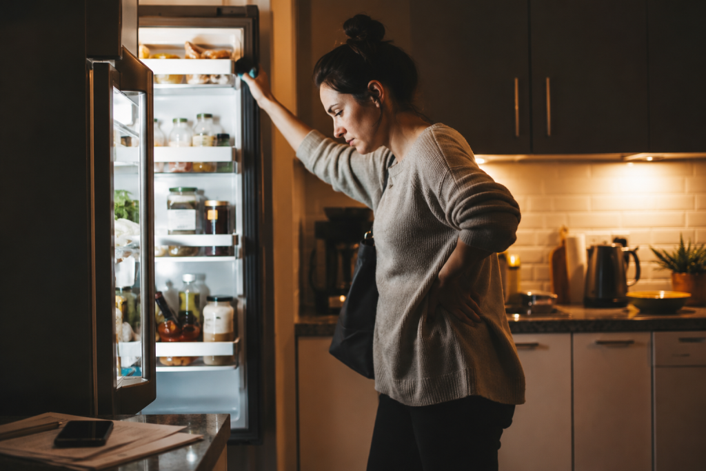 Tired professional standing in the kitchen after work looking into the refrigerator while deciding on a quick and healthy meal.